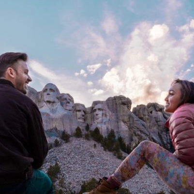Two people sit smiling at each other in front of Mount Rushmore National Memorial