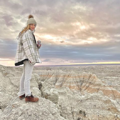 Person dressed in a warm jacket and knit hat stands in Badlands National Park enjoying the muted colors with their morning coffee